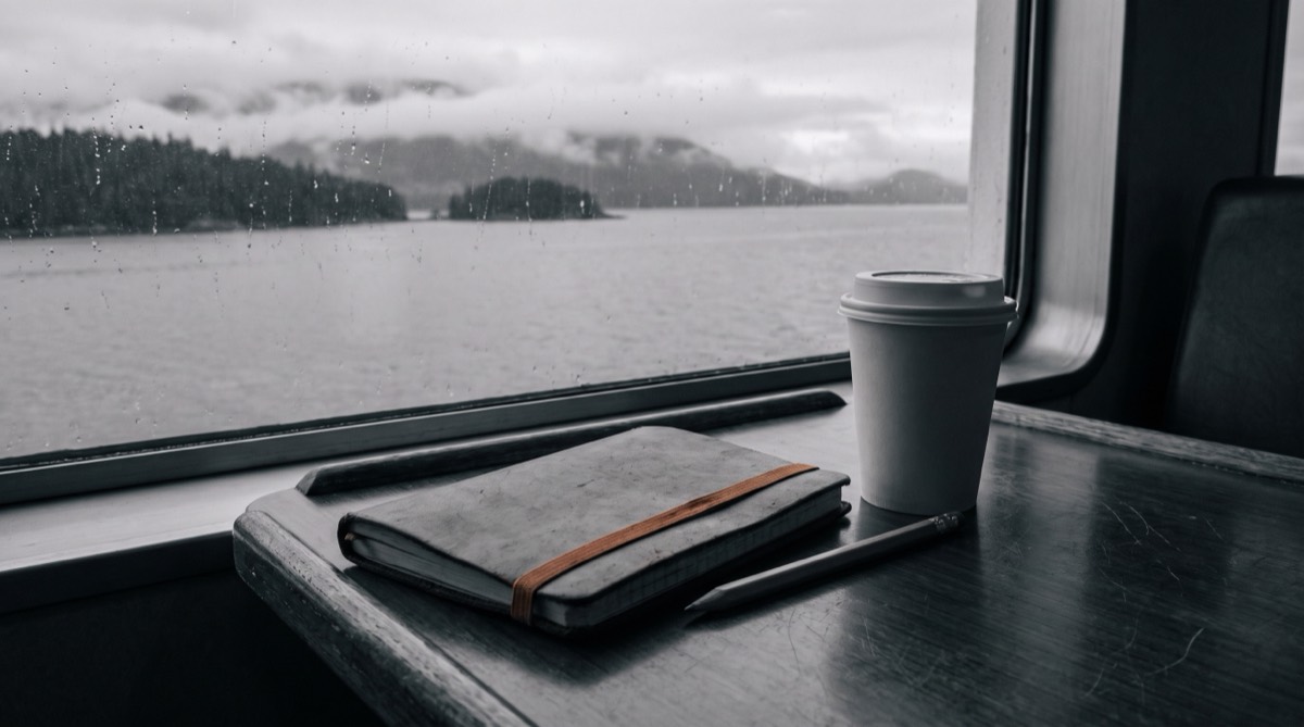 A worn field notebook with a burnt-orange elastic band on a ferry window-seat table, pencil and coffee beside, grey sea and forested islands passing outside.